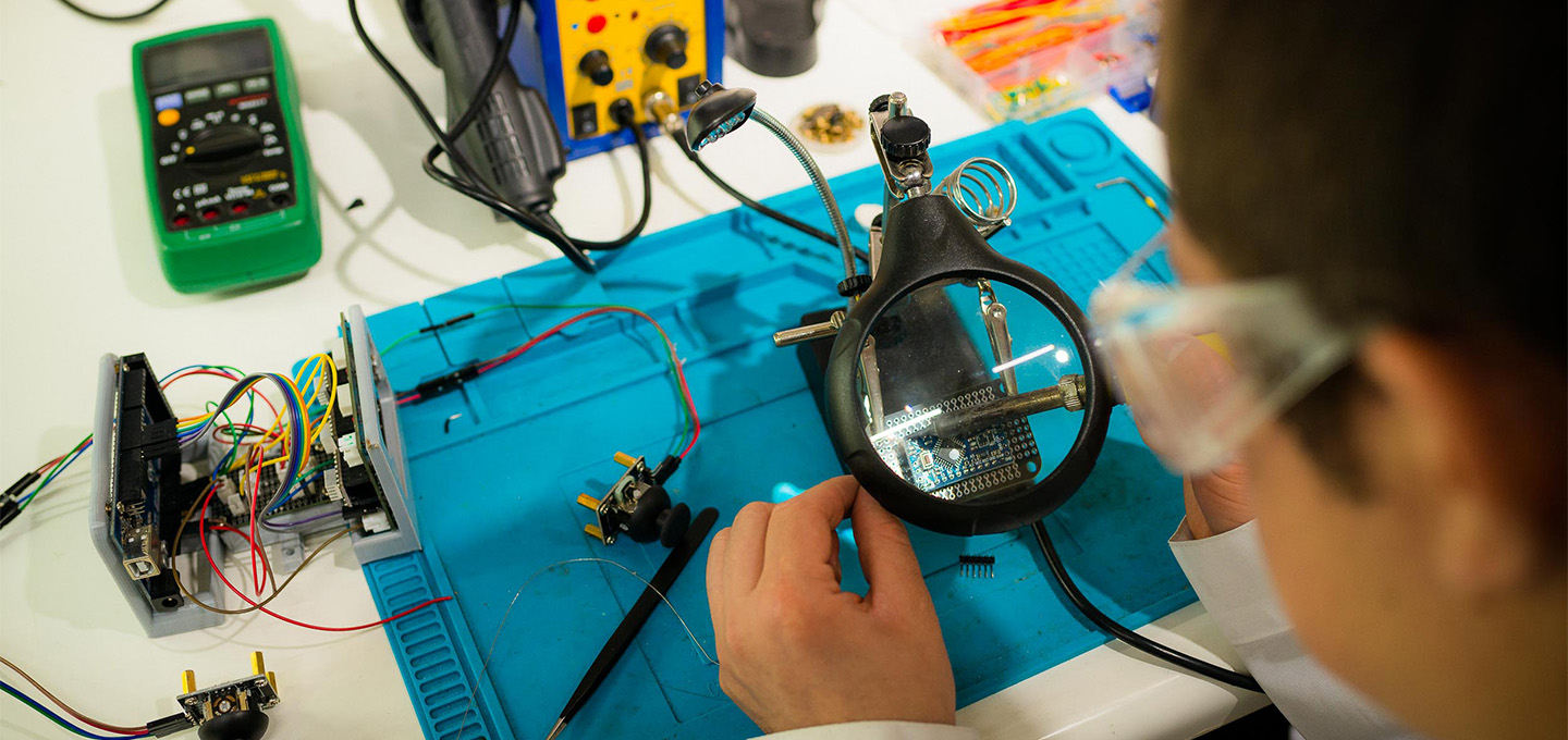 A technician working with electronic components using a magnifying glass and soldering tools.